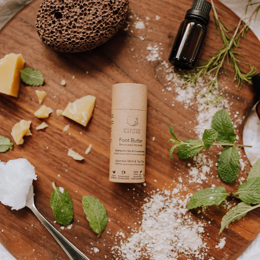 A tube of NATURE Foot Butter placed on a wooden board with mint leaves, salt, soap, a sponge, and an essential oil bottle around it.