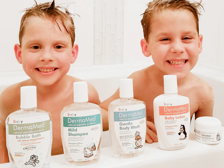 Two smiling young boys sitting in a white bathtub with five DermaMed baby care products lined up in front of them, including Bubble Bath, Mild Shampoo, Gentle Body Wash, Baby Lotion, and a small jar.